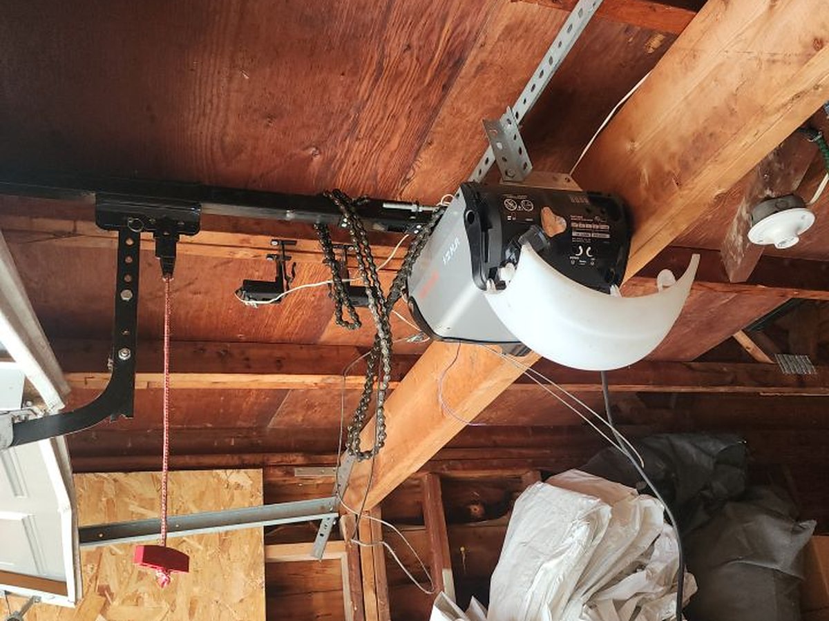 Chain-drive garage door opener mounted to the exposed rafters of a garage, with the rail, chain and emergency release cord visible